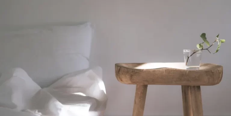 Minimalist bedroom scene with a rustic wooden stool used as a nightstand, holding a small glass of water with a single leafy twig, beside a neatly unmade bed with white linens and pillows in soft natural light.