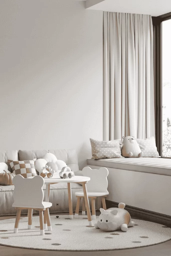 Cozy and modern kids’ playroom with a small round table and two bear-shaped chairs, a neutral-toned sofa with cloud pillows, a soft polka dot rug, and a built-in window seat with plush cushions and stuffed toys, all in calming beige and cream tones.