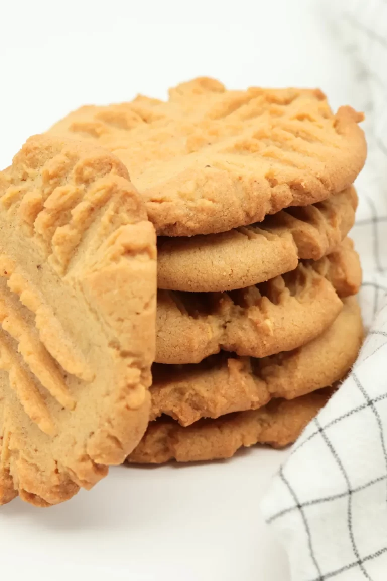 A stack of classic peanut butter cookies with a crisscross pattern, resting next to a cozy white kitchen towel with a black grid design on a light background.
